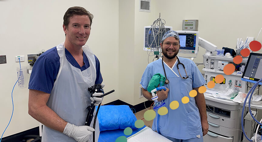 2 men standing in a hospital room, both wearing scrubs and smiling.